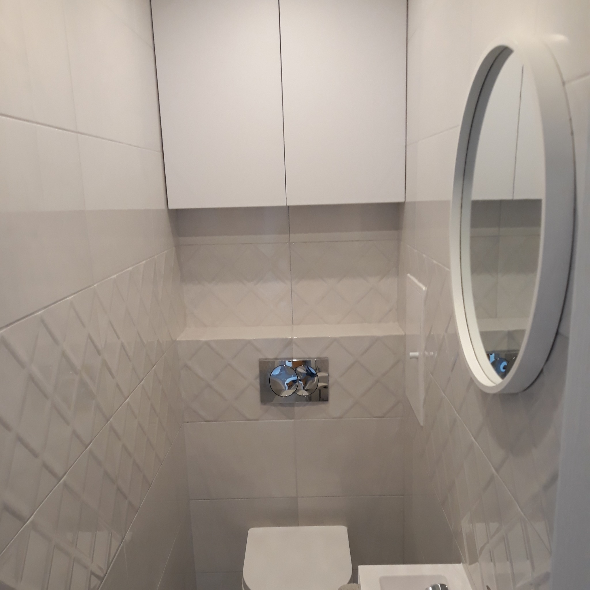 Nowoczesna toaleta z białymi, teksturowanymi płytkami, wiszącą szafką i okrągłym lustrem w minimalistycznej ramie.