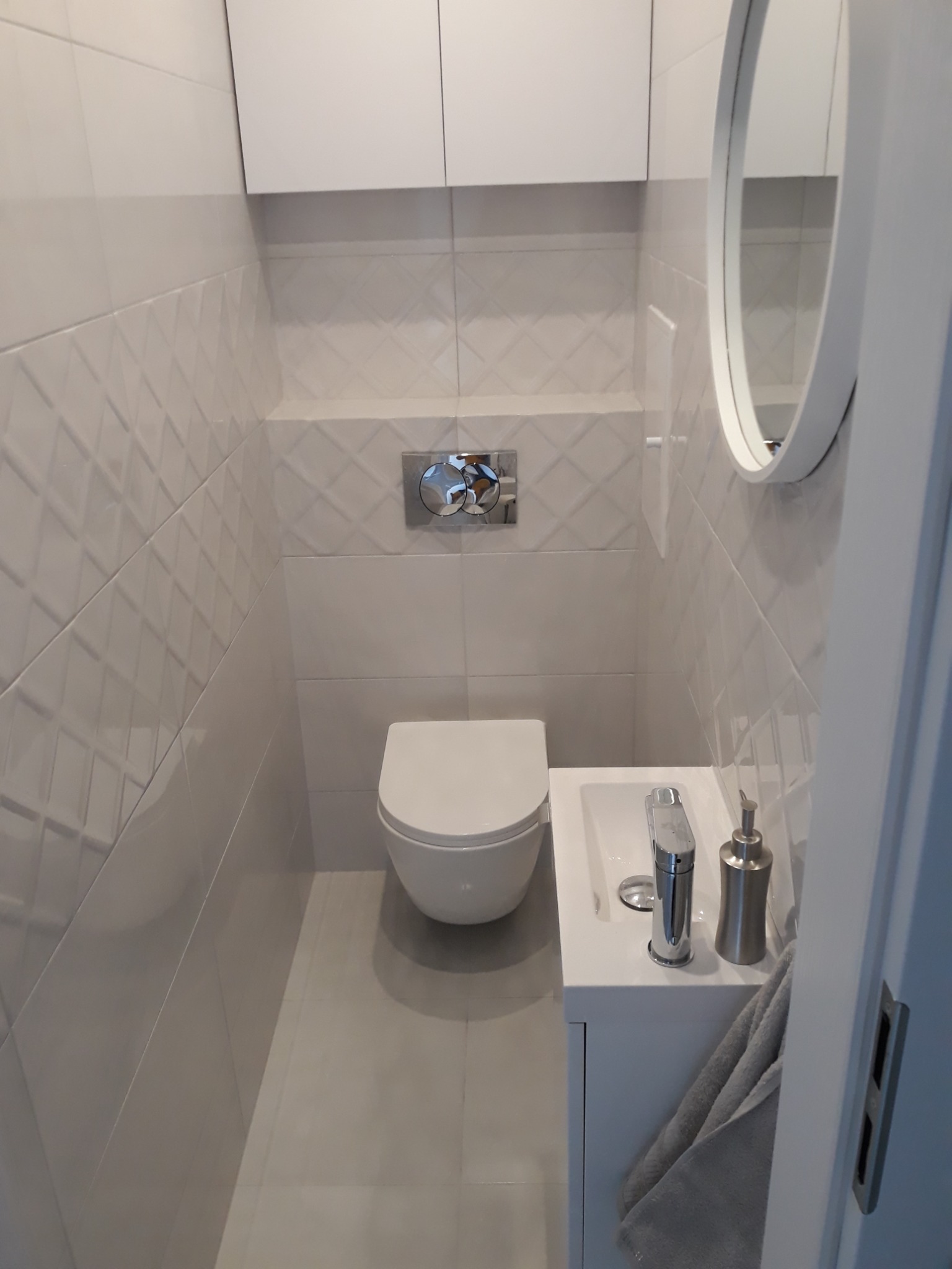 Nowoczesna, minimalistyczna toaleta z białymi, trójwymiarowymi płytkami na ścianach, owalnym lustrem i wiszącą szafką, uzupełniona o srebrną armaturę i dozownik do mydła.