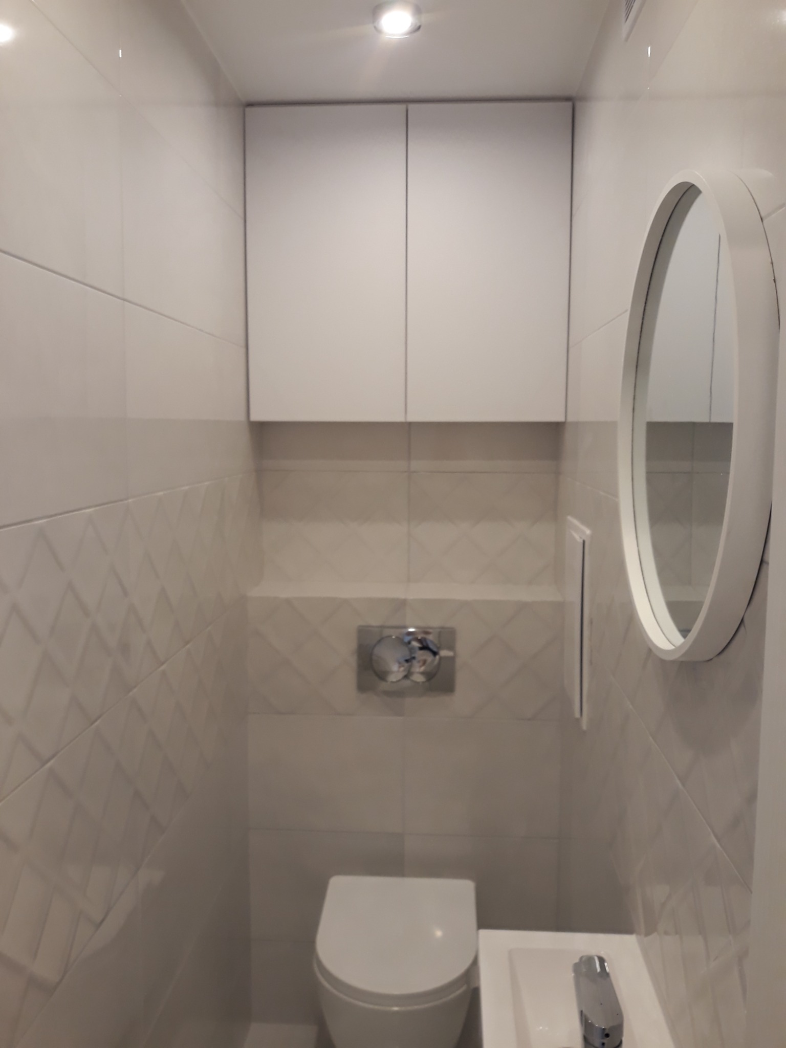 Nowoczesna, minimalistyczna toaleta wykończona białymi płytkami z subtelnym wzorem rombów, uzupełniona okrągłym lustrem i wiszącą szafką.