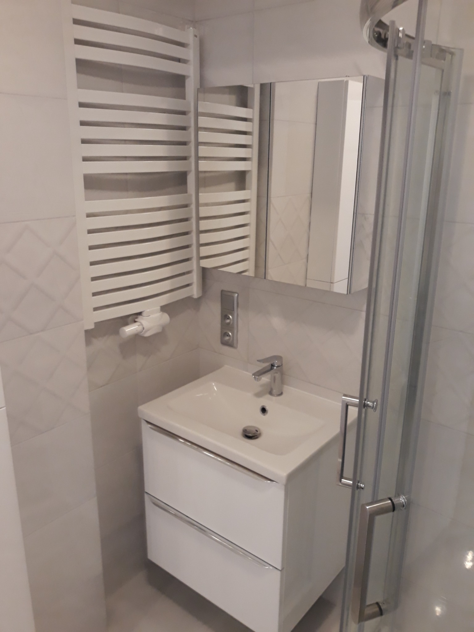 Nowoczesna łazienka z białą umywalką z szafką, grzejnikiem drabinkowym i przeszkloną kabiną prysznicową.