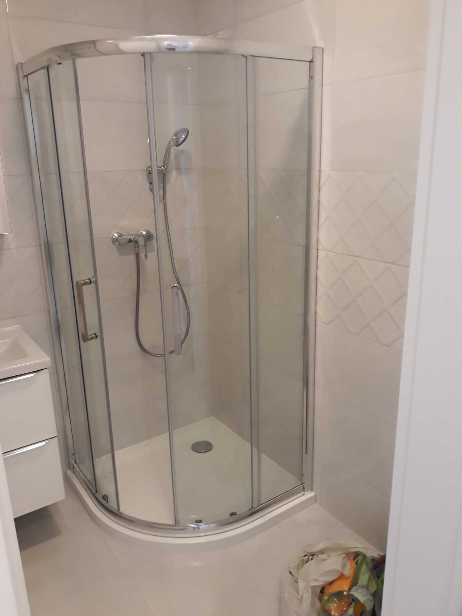 Narożna kabina prysznicowa z chromowanymi profilami i przezroczystymi szybami, zamontowana w łazience z jasnymi płytkami w romby, widoczna bateria i słuchawka prysznicowa, obok umywalka z szafką...