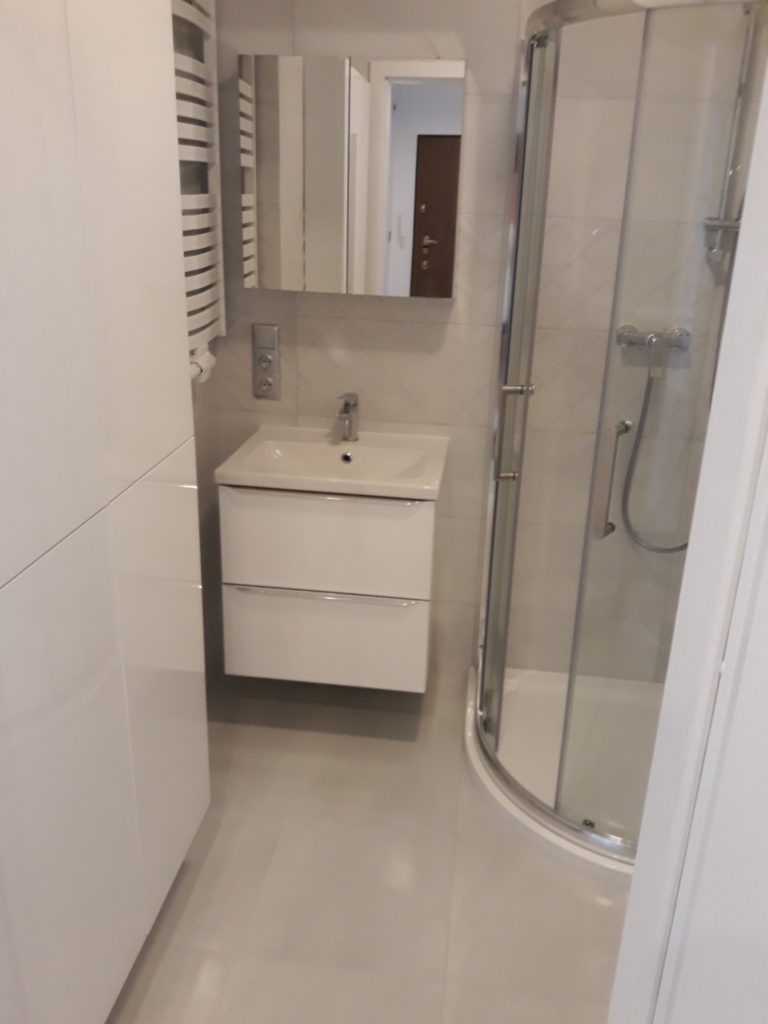 Nowoczesna łazienka z białą szafką umywalkową, lustrem, grzejnikiem drabinkowym i kabiną prysznicową z przezroczystego szkła, wykończona w jasnych odcieniach beżu i bieli.