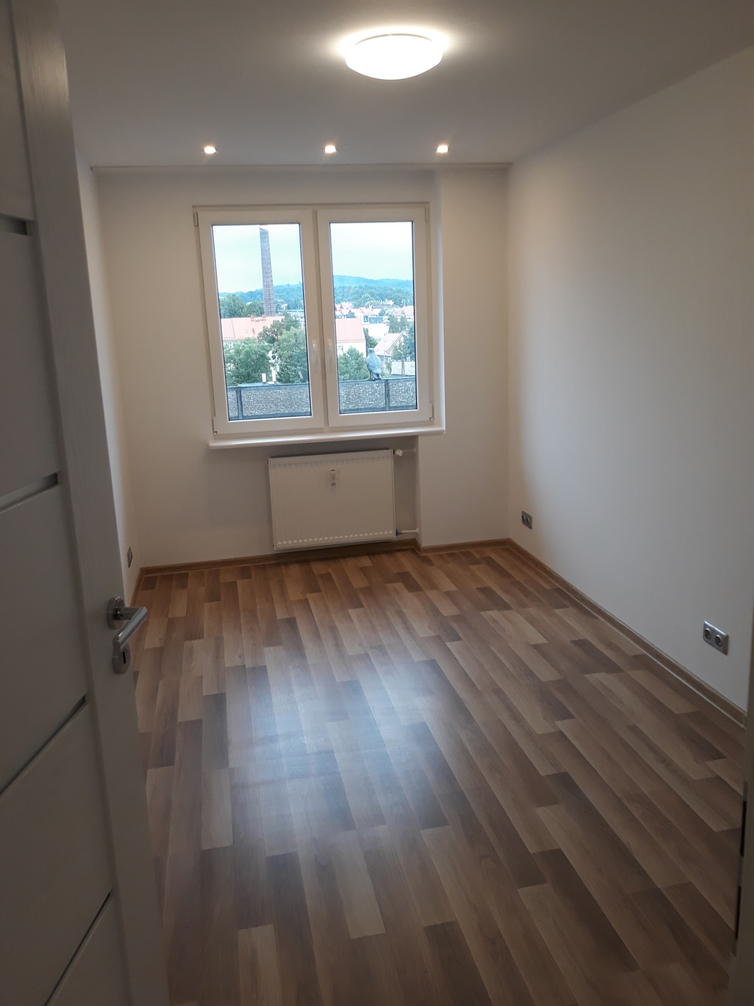 Jasne wnętrze pokoju z drewnopodobną podłogą, białymi ścianami i oknem z widokiem na miasto, oświetlone światłem z sufitowych halogenów.