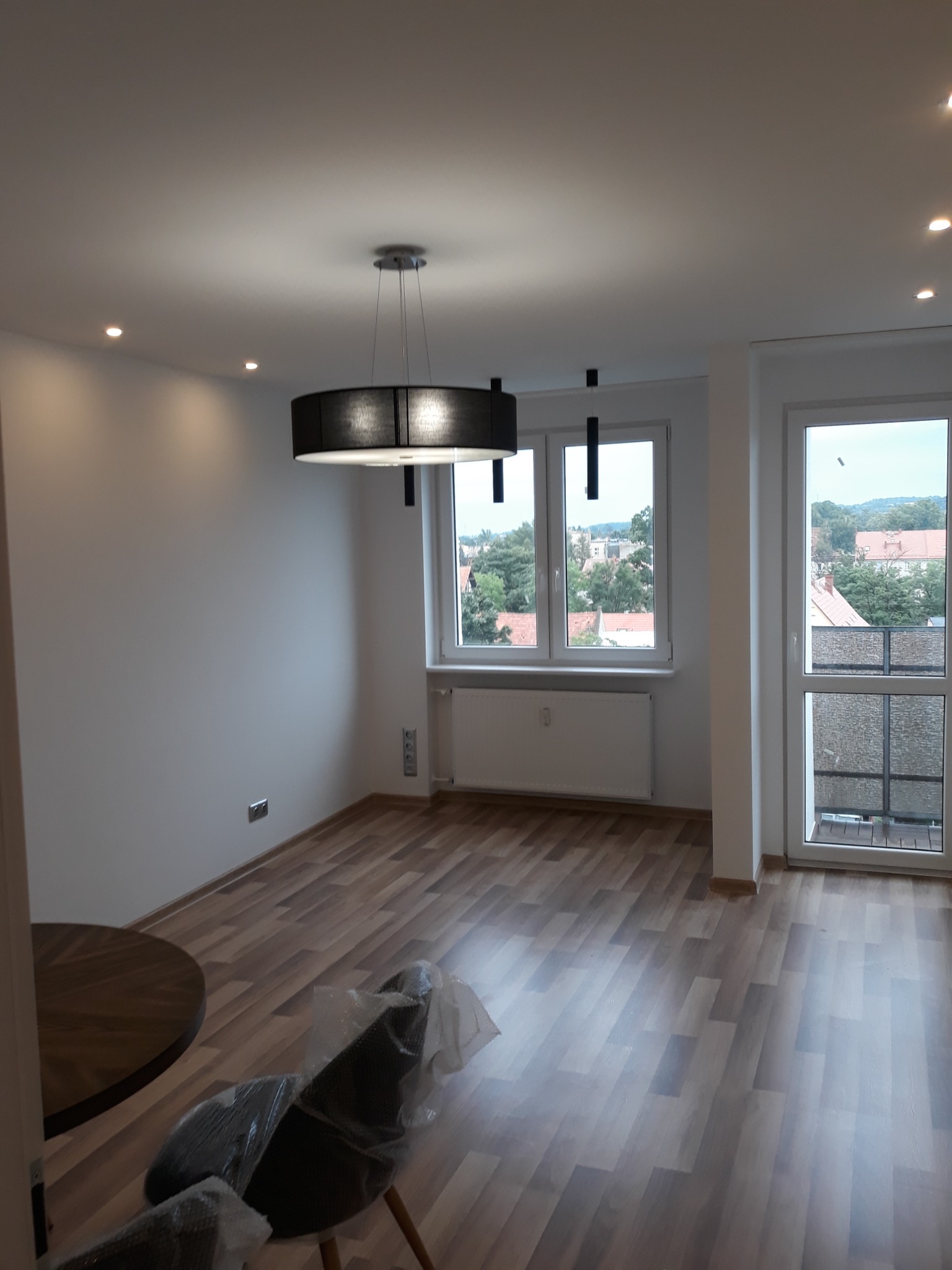 Nowe panele podłogowe w jasnym, przestronnym pokoju z dużymi oknami i nowoczesnym oświetleniem sufitowym, krzesła zabezpieczone folią.