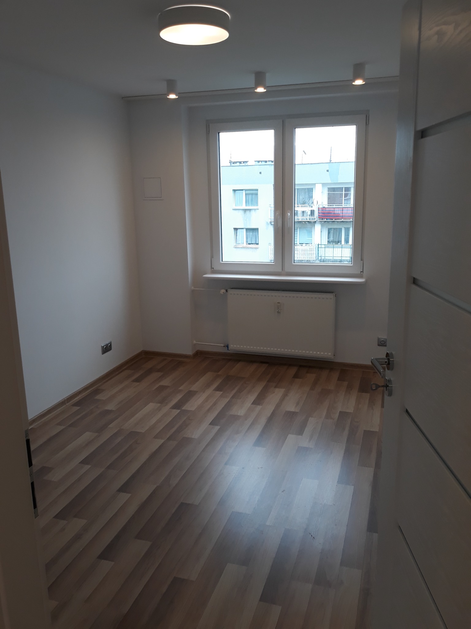 Pusty pokój z panelami podłogowymi ułożonymi w jodełkę, białe ściany, okno z widokiem na budynek mieszkalny i grzejnik pod oknem, oświetlenie sufitowe.