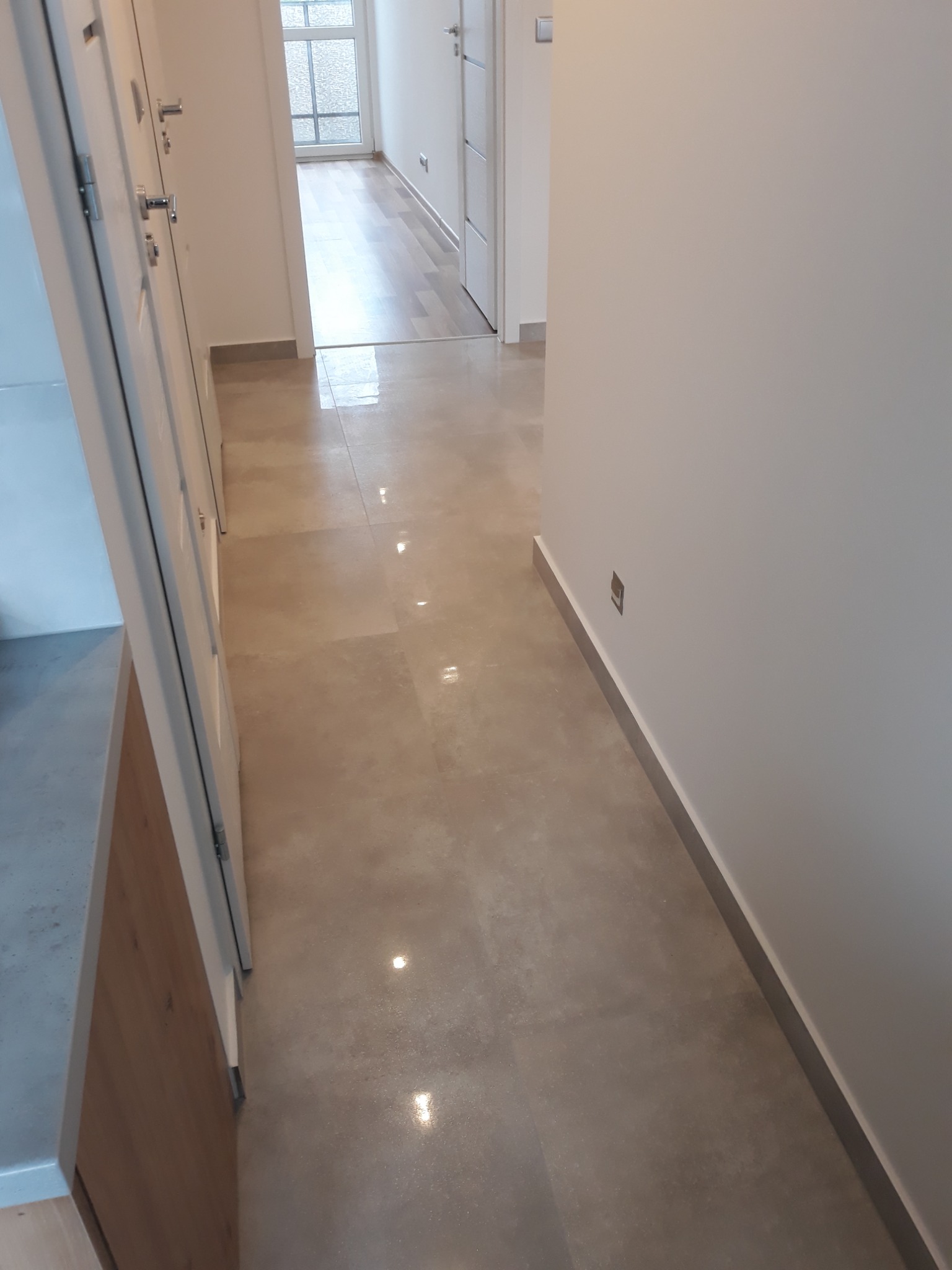 Beżowa, błyszcząca podłoga w korytarzu z widoczną listwą przypodłogową i fragmentem szafki z drewnianym frontem.