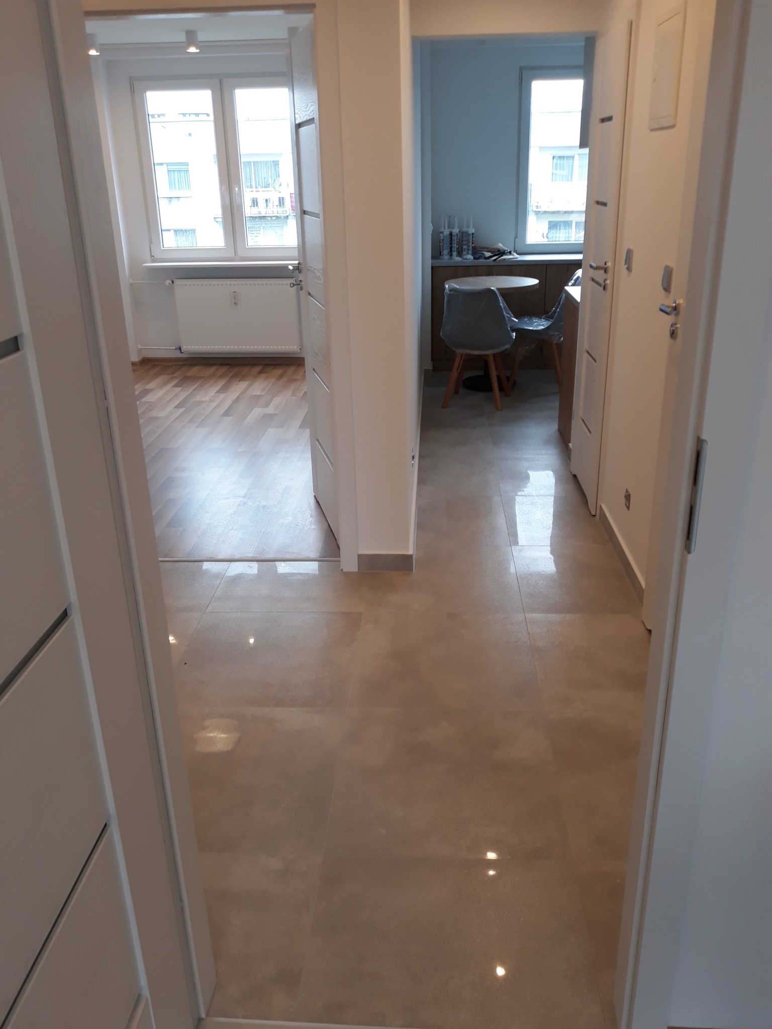 Beżowe płytki podłogowe z połyskiem w korytarzu, widok z perspektywy drzwi do salonu z drewnianą podłogą i kuchni z szarą krzesłem i stolikiem