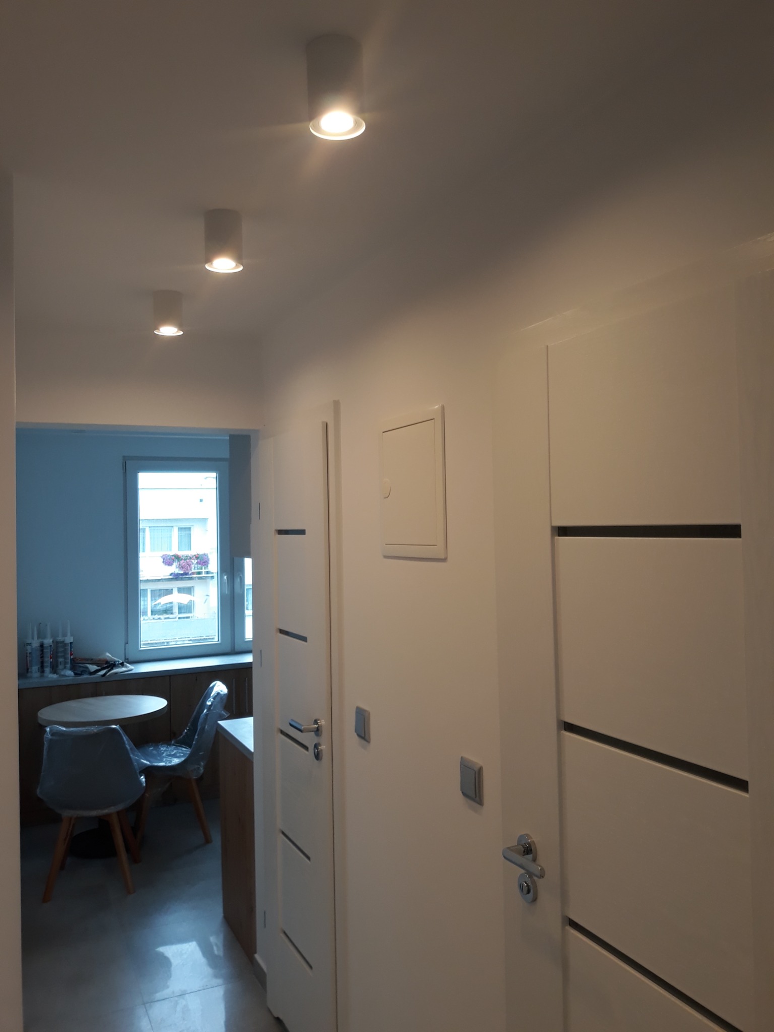 Nowoczesny korytarz z białymi ścianami i drzwiami z poziomymi wstawkami, oświetlony trzema okrągłymi lampami sufitowymi. W tle widoczny fragment kuchni z oknem, stolikiem i krzesłami.