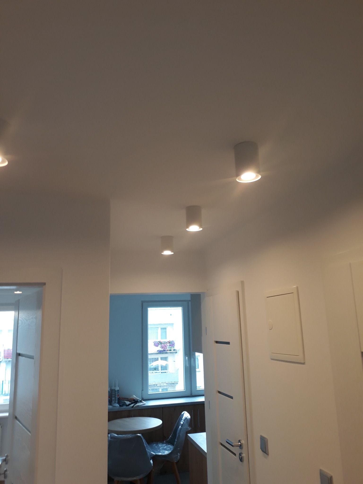 Nowoczesne, białe wnętrze mieszkania z trzema okrągłymi lampami sufitowymi typu downlight, widok na białe ściany, drzwi wewnętrzne z poziomymi frezami i fragment jadalni z okrągłym stołem...