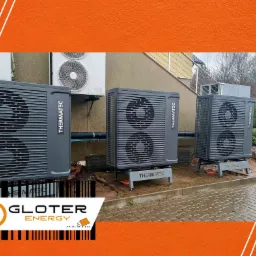 Trzy szare jednostki zewnętrzne pomp ciepła Thermatec ustawione obok siebie na betonowej powierzchni, z widocznymi elementami instalacji i budynkiem w tle. Na pierwszym planie logo firmy Gloter...