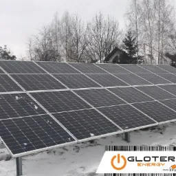 Instalacja fotowoltaiczna na śniegu, widoczne panele słoneczne na metalowych wspornikach, w tle drzewa i fragment budynku, pochmurne niebo.