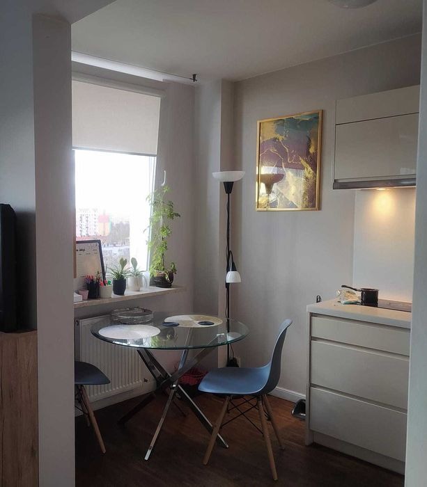 Jasne wnętrze kuchni z okrągłym, szklanym stołem, dwoma krzesłami w stylu skandynawskim, minimalistyczną lampą podłogową i abstrakcyjnym obrazem w złotej ramie na ścianie, widok zza filaru.