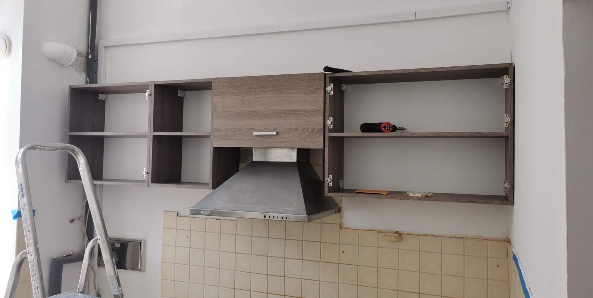 Montaż szafek kuchennych wiszących z drewnopodobnym fornirem, widoczny okap, stara glazura i drabina aluminiowa.