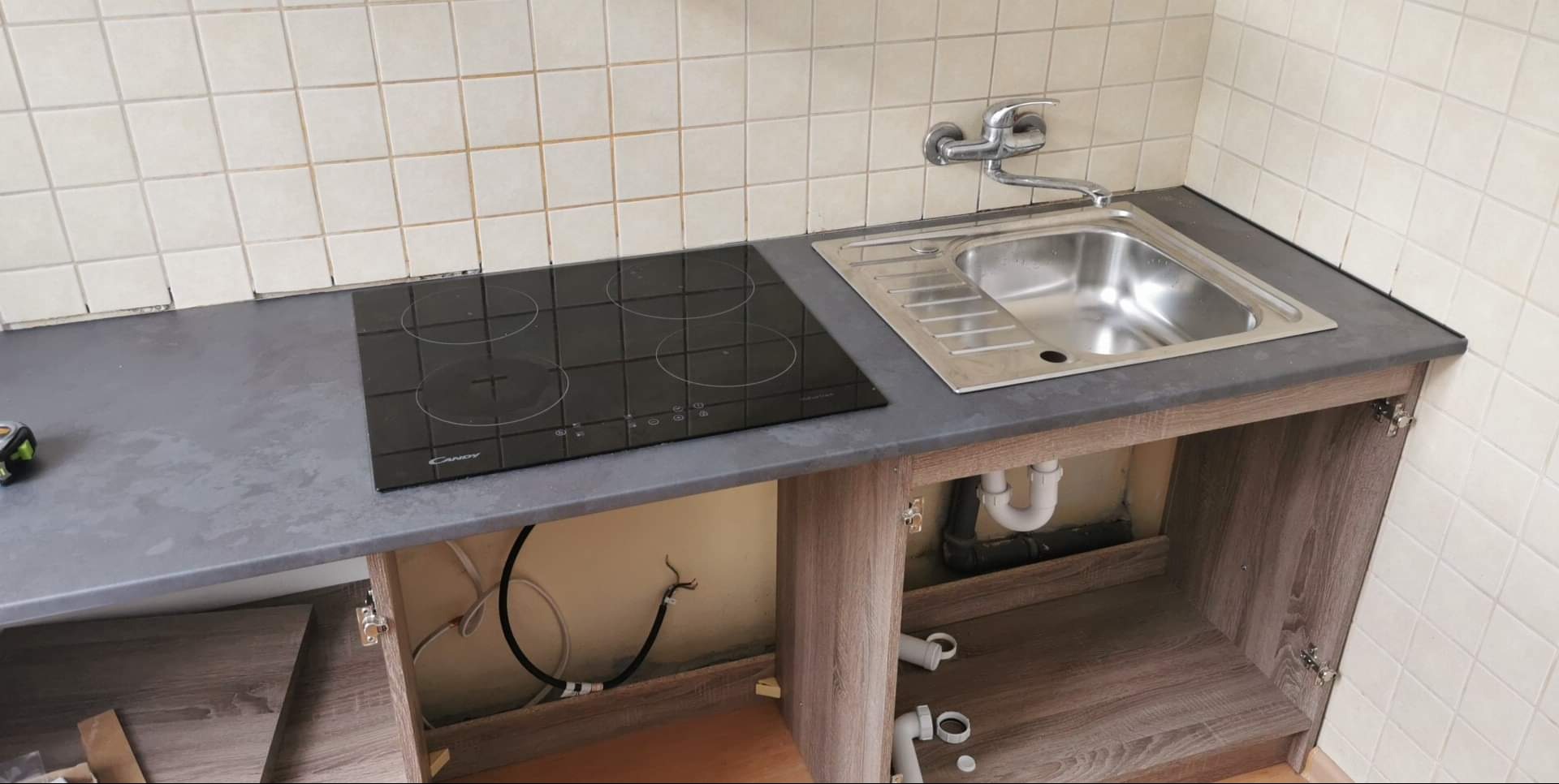 Nowa płyta indukcyjna i zlewozmywak zamontowane na szarym blacie kuchennym w trakcie prac wykończeniowych, widoczne niedociągnięcia przy łączeniu płytek, otwarte szafki pod zlewem z widoczną...