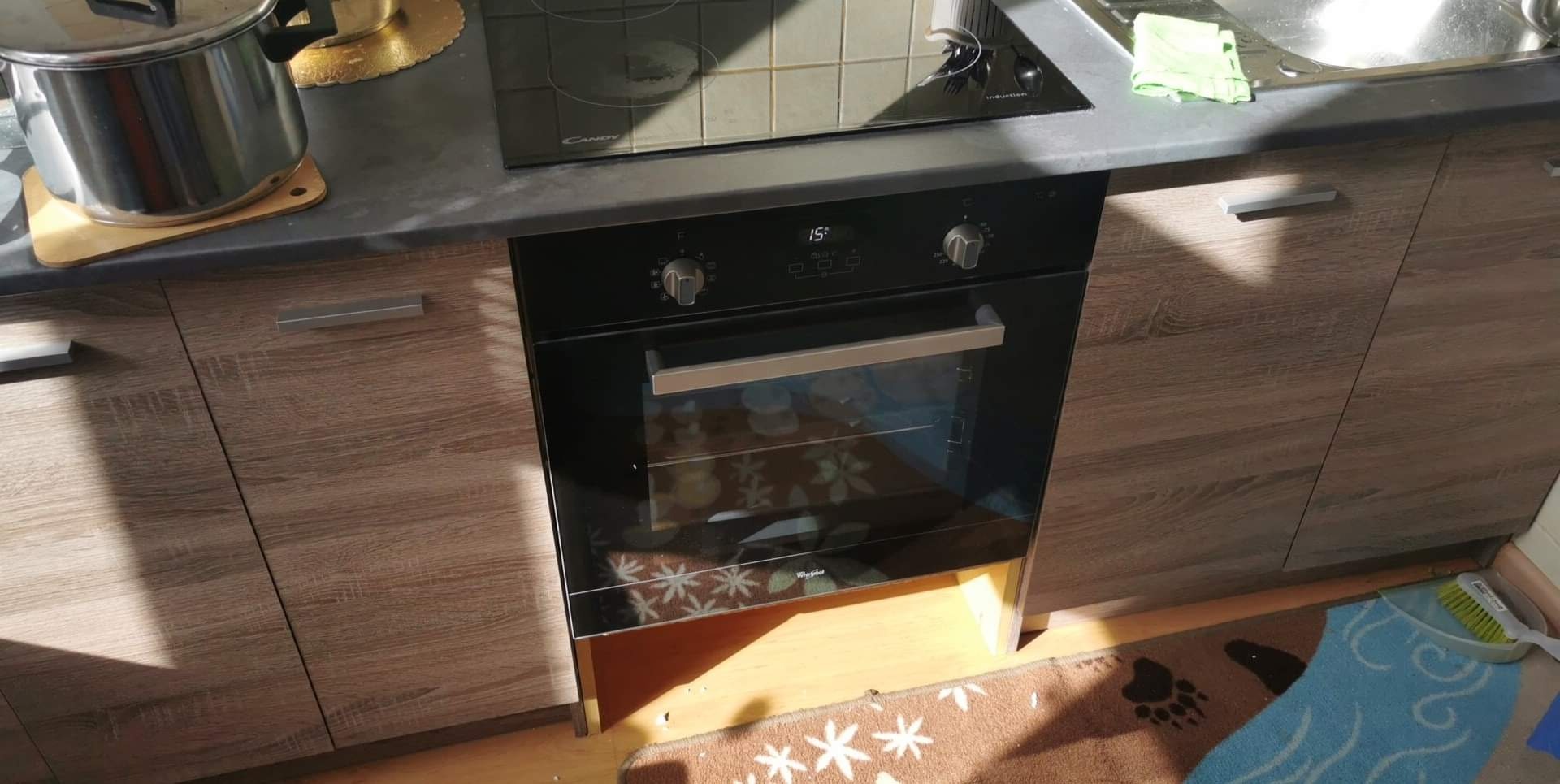 Kuchnia z zabudową drewnopodobną, czarnym blatem, płytą indukcyjną i piekarnikiem Whirlpool, widoczny fragment dywanika pod szafkami.