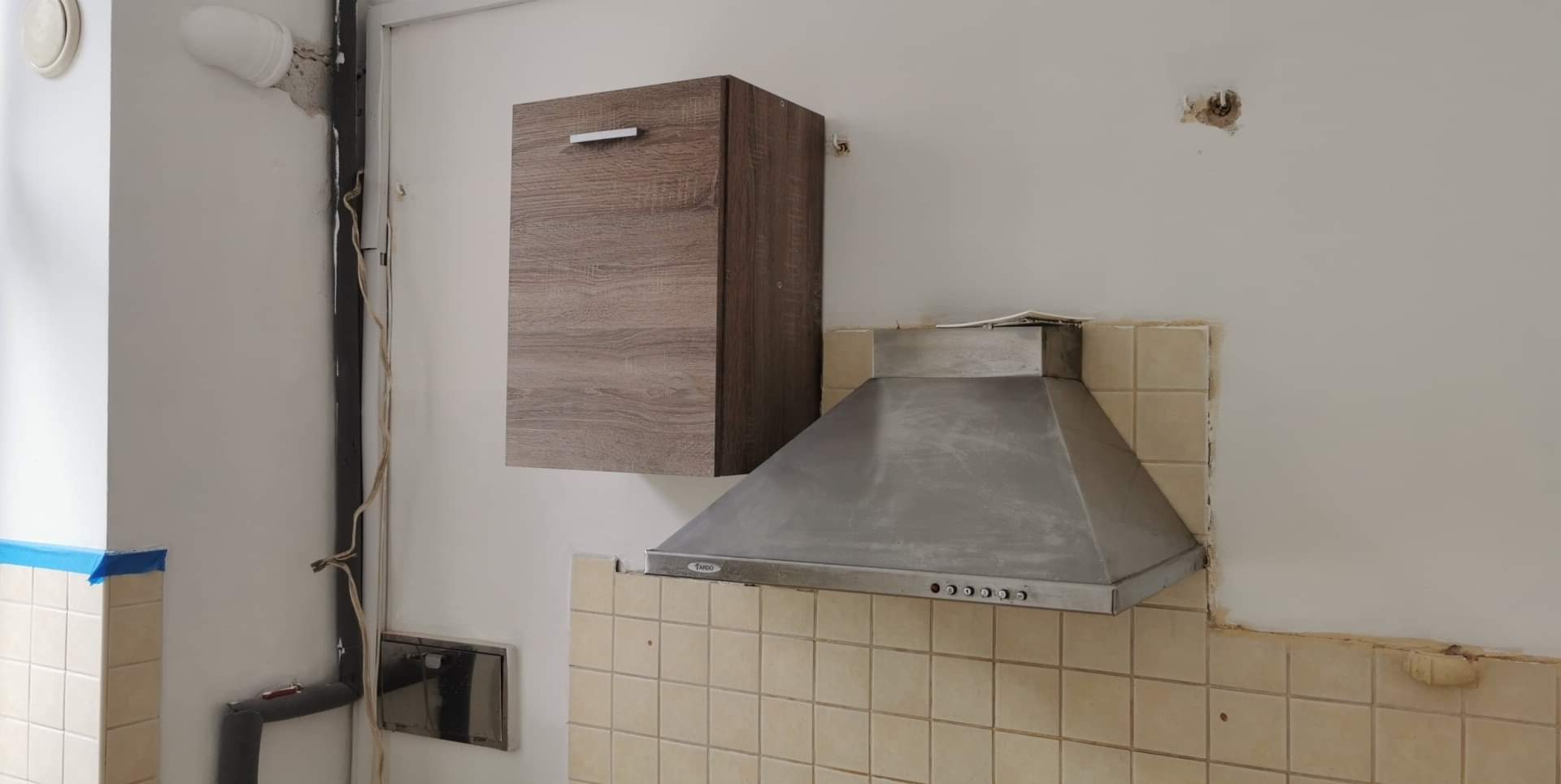 Kuchnia w trakcie prac wykończeniowych: wisząca szafka, okap nad płytkami, ślady po instalacjach na ścianie, widoczne przygotowania do malowania.