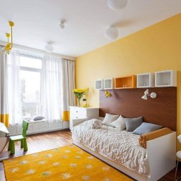 Renovation sp z o.o. - Jasny pokój dziecięcy z żółtą ścianą, drewnianym zagłówkiem łóżka, białymi półkami, wzorzystym dywanem i naturalnym światłem wpadającym przez okno.