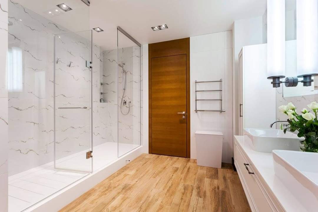 Nowoczesna łazienka z kabiną prysznicową typu walk-in, drewnianą podłogą i białymi elementami wyposażenia, w tym podwójną umywalką i grzejnikiem drabinkowym.