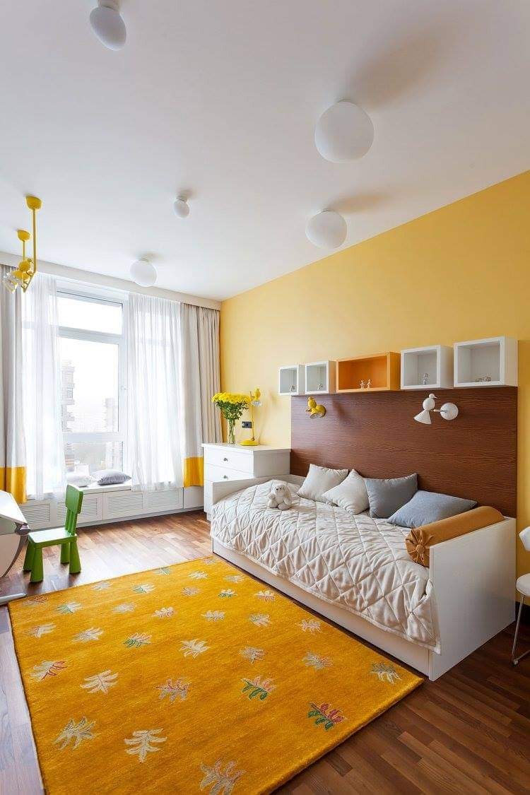 Jasny pokój dziecięcy z żółtą ścianą, drewnianym zagłówkiem łóżka, białymi półkami, wzorzystym dywanem i naturalnym światłem wpadającym przez okno.