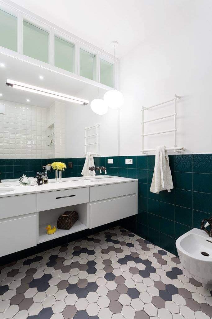 Nowoczesna łazienka z białą armaturą, szafką pod umywalką, ciemnozielonymi płytkami i heksagonalną mozaiką na podłodze. Na blacie flakon z żółtymi kwiatami i kosmetyki.