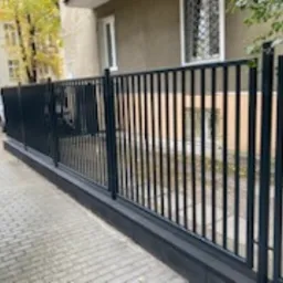 Ciemnoszare, metalowe ogrodzenie panelowe z furtką na tle chodnika z kostki i budynku mieszkalnego z widocznymi oknami.