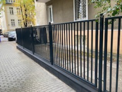 Ciemnoszare, metalowe ogrodzenie panelowe z furtką na tle chodnika z kostki i budynku mieszkalnego z widocznymi oknami.