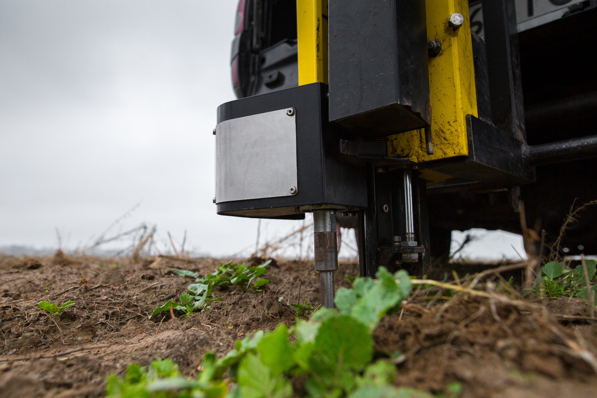 Detail ujęcia wiertnicy studni głębinowej z żółto-czarną konstrukcją, wbijającej się w ziemię z młodymi roślinami, pochmurne tło.