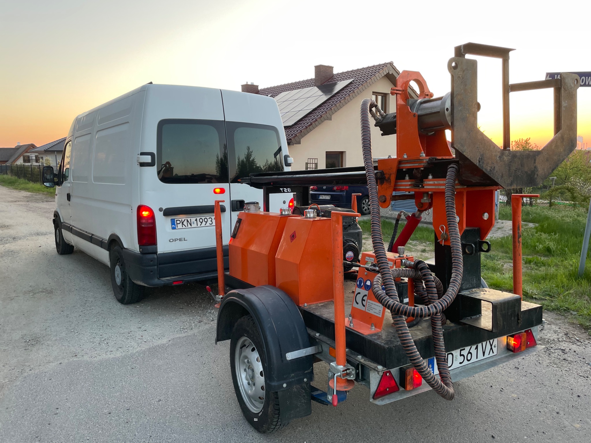 Wiertnica zamontowana na przyczepie, podłączona do białego samochodu dostawczego Opel, na tle domu z panelami słonecznymi i zachodzącego słońca.