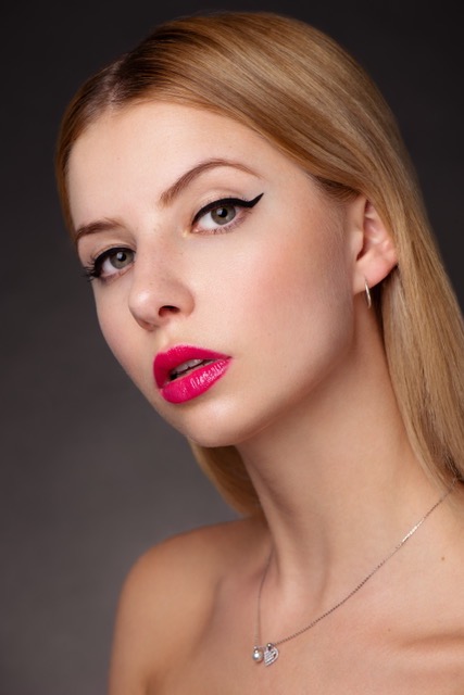 Portret młodej kobiety z precyzyjną kreską eyelinerem i intensywnie różową szminką, delikatny srebrny naszyjnik z zawieszką w kształcie serca.