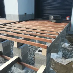 Konstrukcja tarasu z drewnianych legarów na betonowych słupach, widoczne elementy montażowe i podłoże z kamieni. Etap budowy tarasu przy budynku.