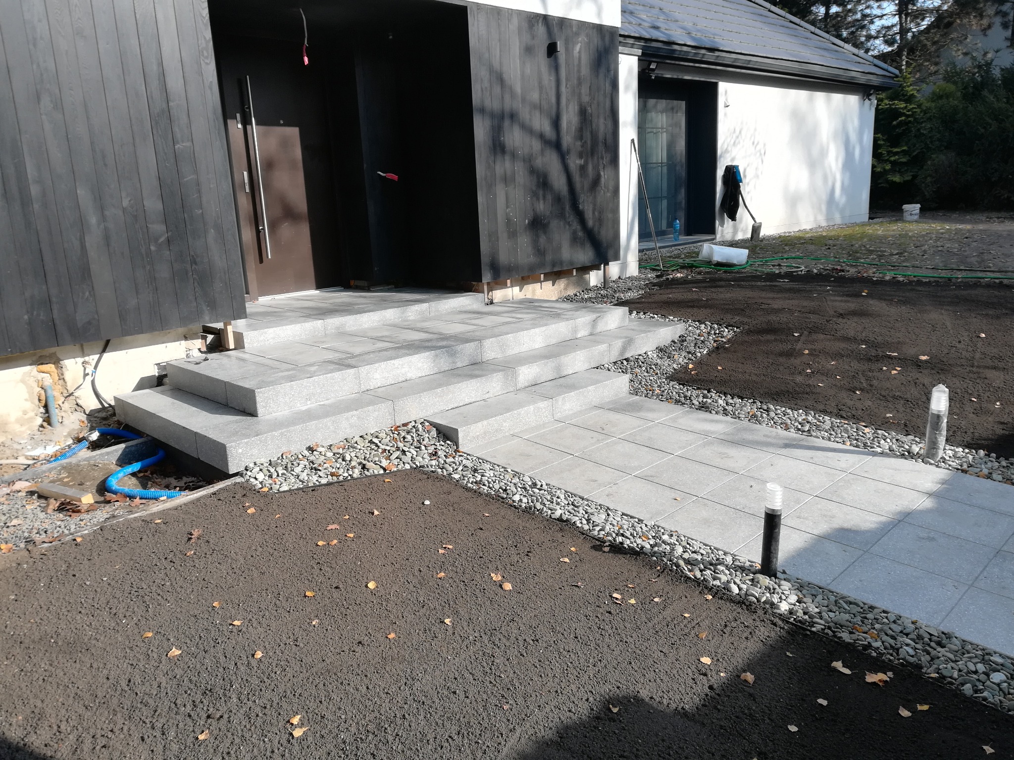 Nowoczesne schody i chodnik z jasnoszarej kostki granitowej prowadzące do domu z elementami czarnego drewna, świeżo przygotowany grunt pod trawnik, oświetlenie.