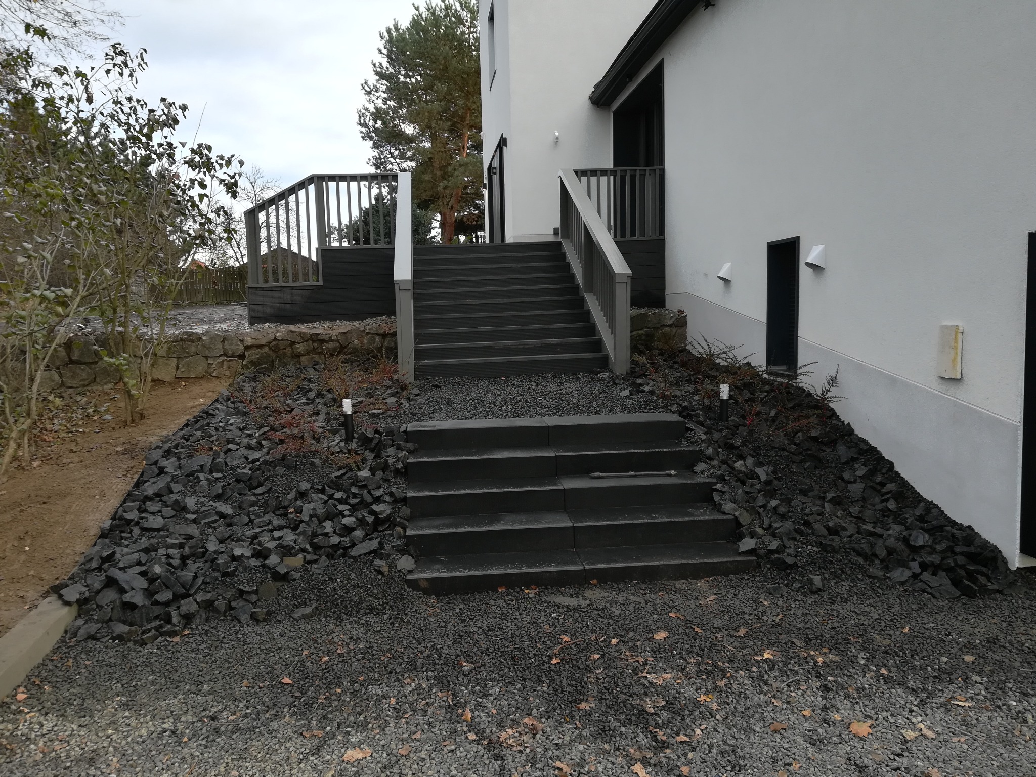 Nowoczesne schody zewnętrzne z ciemnego drewna kompozytowego prowadzące do tarasu, otoczone dekoracyjnym kamieniem i żwirem w minimalistycznym stylu.