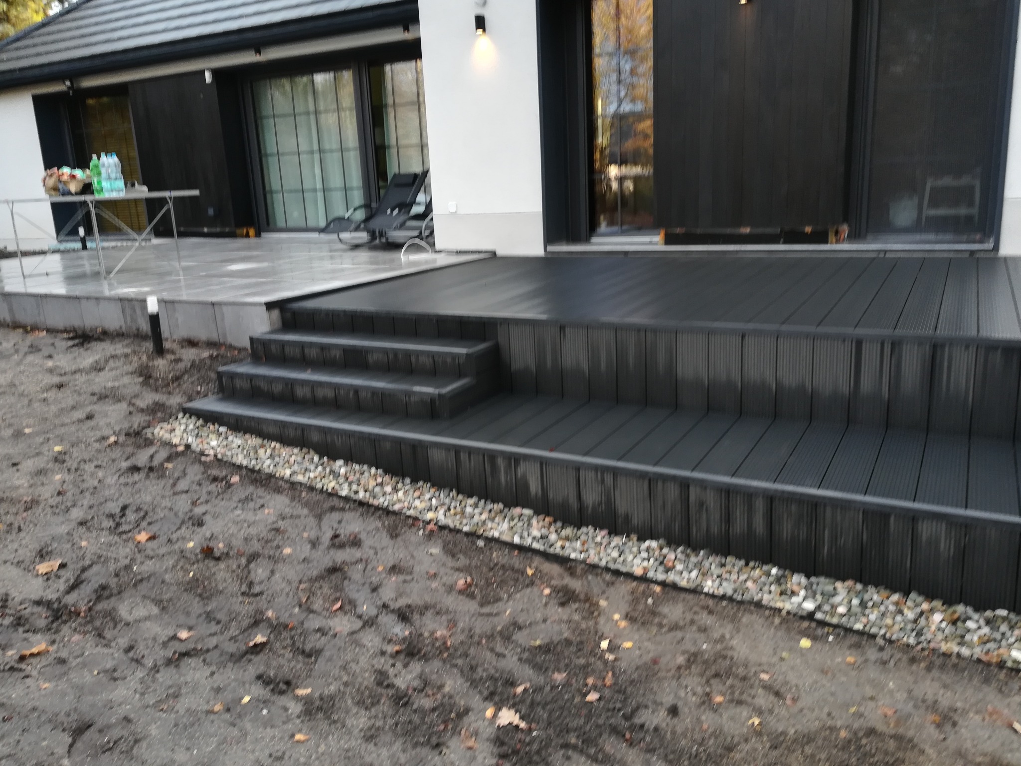 Nowoczesny taras kompozytowy w odcieniach szarości, z niskimi schodami i drobnym żwirem u podstawy. W tle widoczny fragment domu z dużymi oknami i tarasem.