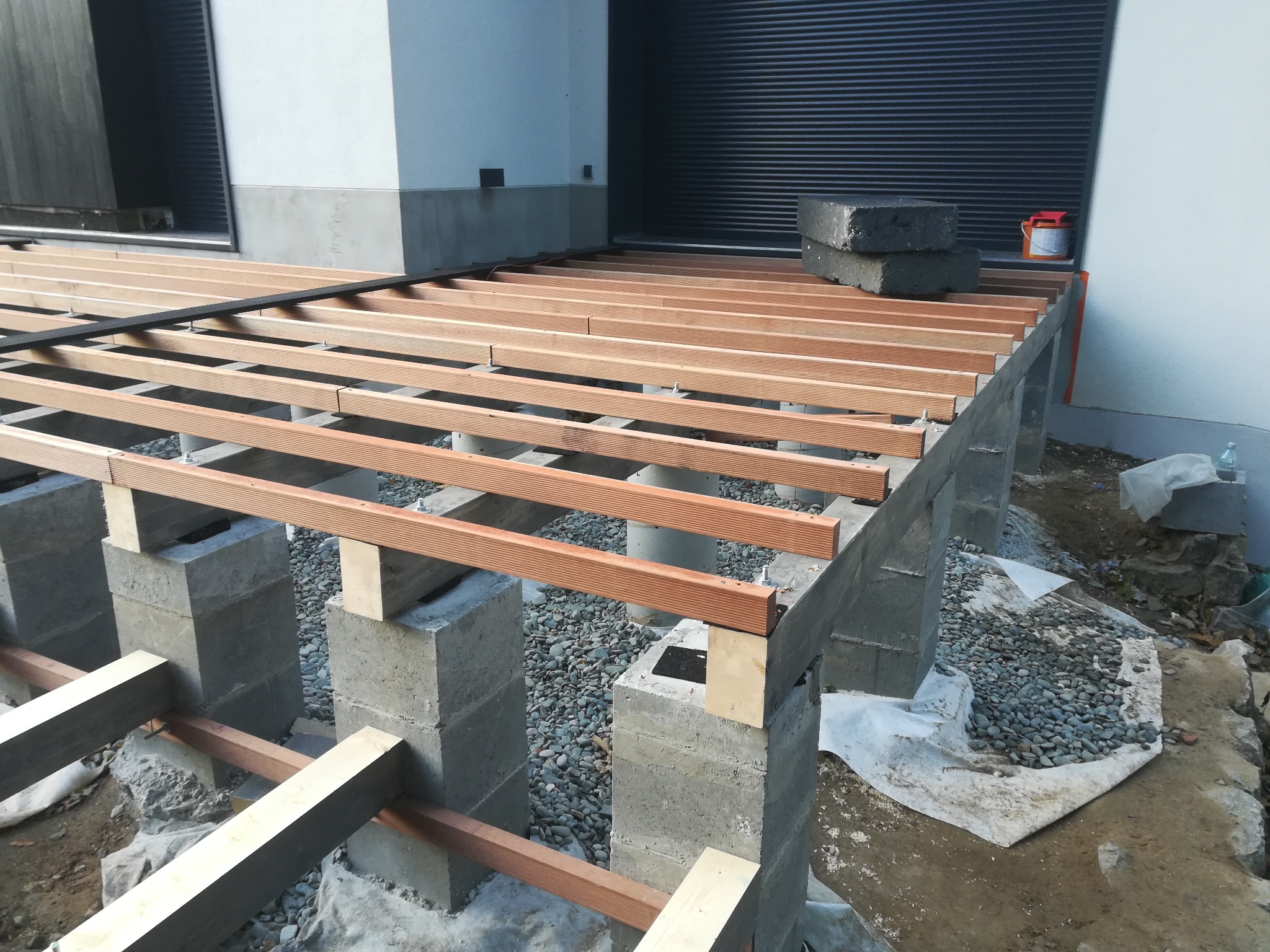 Konstrukcja tarasu z drewnianych legarów na betonowych słupach, widoczne elementy montażowe i podłoże z kamieni. Etap budowy tarasu przy budynku.