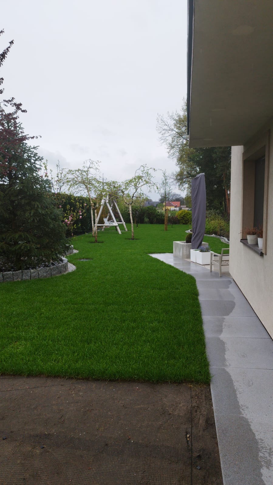Zadbany trawnik z młodymi drzewkami i huśtawką w ogrodzie. Szary chodnik prowadzi wzdłuż domu. Widoczne rabaty z kamienia i rośliny.