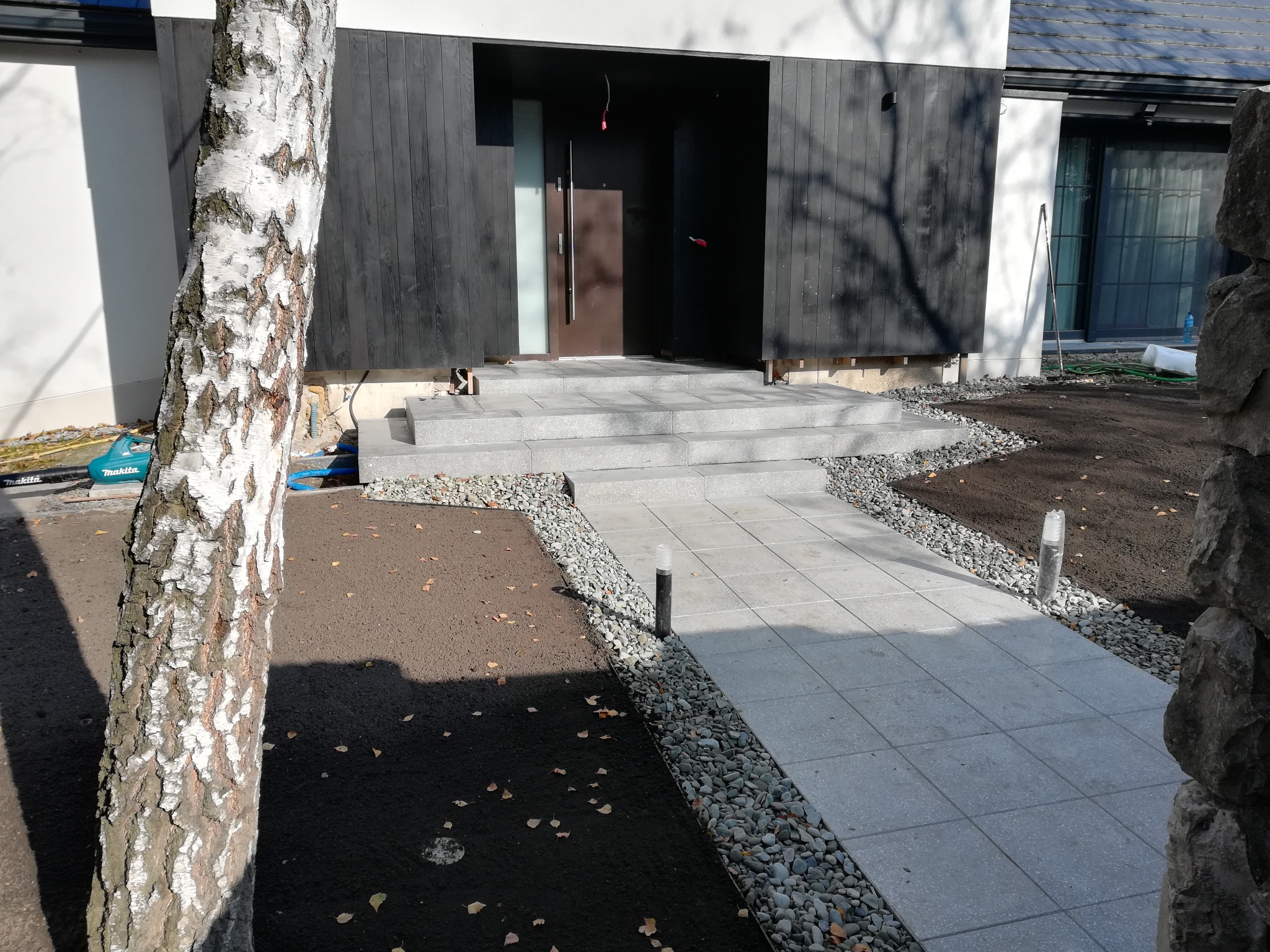 Nowoczesne wejście do domu z szarej kostki, obrzeża z kamyków. Po lewej brzoza, po prawej kamienny element. Minimalistyczny design z elementami naturalnymi.