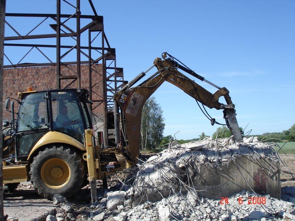Żółta koparko-ładowarka CAT kruszy betonowy fundament z wystającymi prętami zbrojeniowymi na tle konstrukcji stalowej i ceglanego budynku w słoneczny dzień.