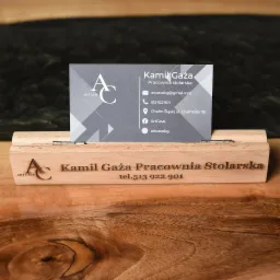 Wizytówka pracowni stolarskiej Kamil Gaża umieszczona w drewnianym stojaku z wygrawerowaną nazwą i numerem telefonu, na tle drewnianego blatu.