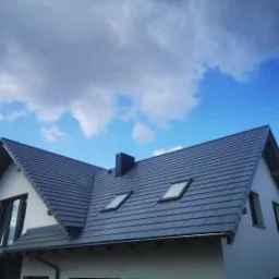 Nowoczesny dach z ciemnej dachówki ceramicznej z dwoma oknami połaciowymi i kominem na tle błękitnego nieba z chmurami.