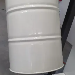 Biała, metalowa beczka po malowaniu proszkowym, zawieszona na konstrukcji w warsztacie, z widocznymi śladami użytkowania na platformie pod nią.