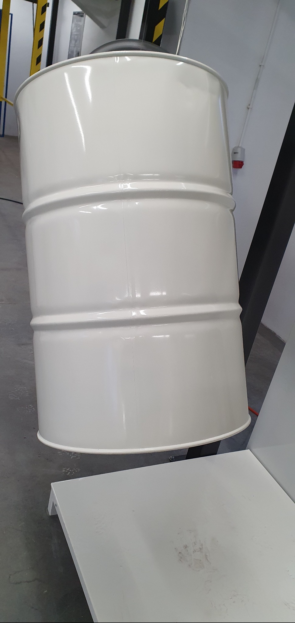 Biała, metalowa beczka po malowaniu proszkowym, zawieszona na konstrukcji w warsztacie, z widocznymi śladami użytkowania na platformie pod nią.