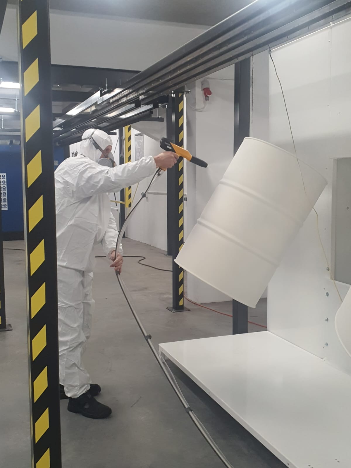 Pracownik w kombinezonie ochronnym aplikuje farbę na metalową beczkę za pomocą pistoletu natryskowego w hali produkcyjnej.