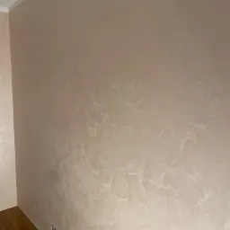 Beżowa ściana z dekoracyjną strukturą, widoczne gniazdka elektryczne, fragment drewnianej podłogi i listwa przypodłogowa.