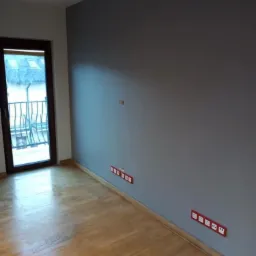 Pomalowane na szaro wnętrze pokoju z drewnianą podłogą i zamontowanymi gniazdkami elektrycznymi w czerwonych ramkach przy ścianie, widok na balkon.