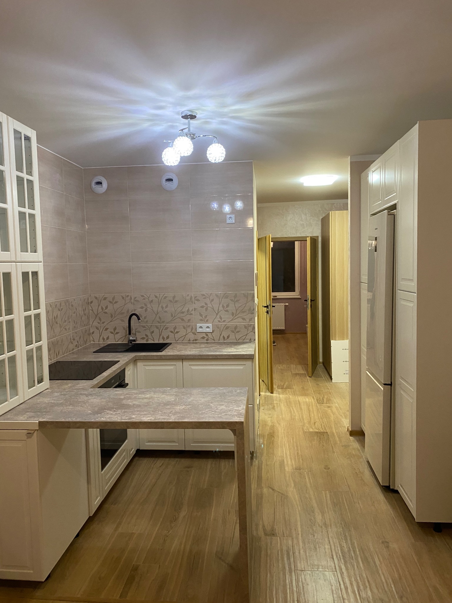 Jasna kuchnia z drewnopodobną podłogą, beżowymi płytkami z motywem liści nad blatem i białymi szafkami z przeszklonymi witrynami, widok na otwarte drzwi do kolejnego pomieszczenia.