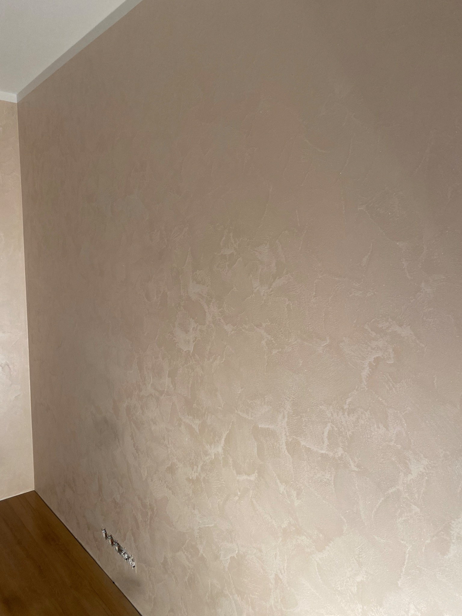 Beżowa ściana z dekoracyjną strukturą, widoczne gniazdka elektryczne, fragment drewnianej podłogi i listwa przypodłogowa.