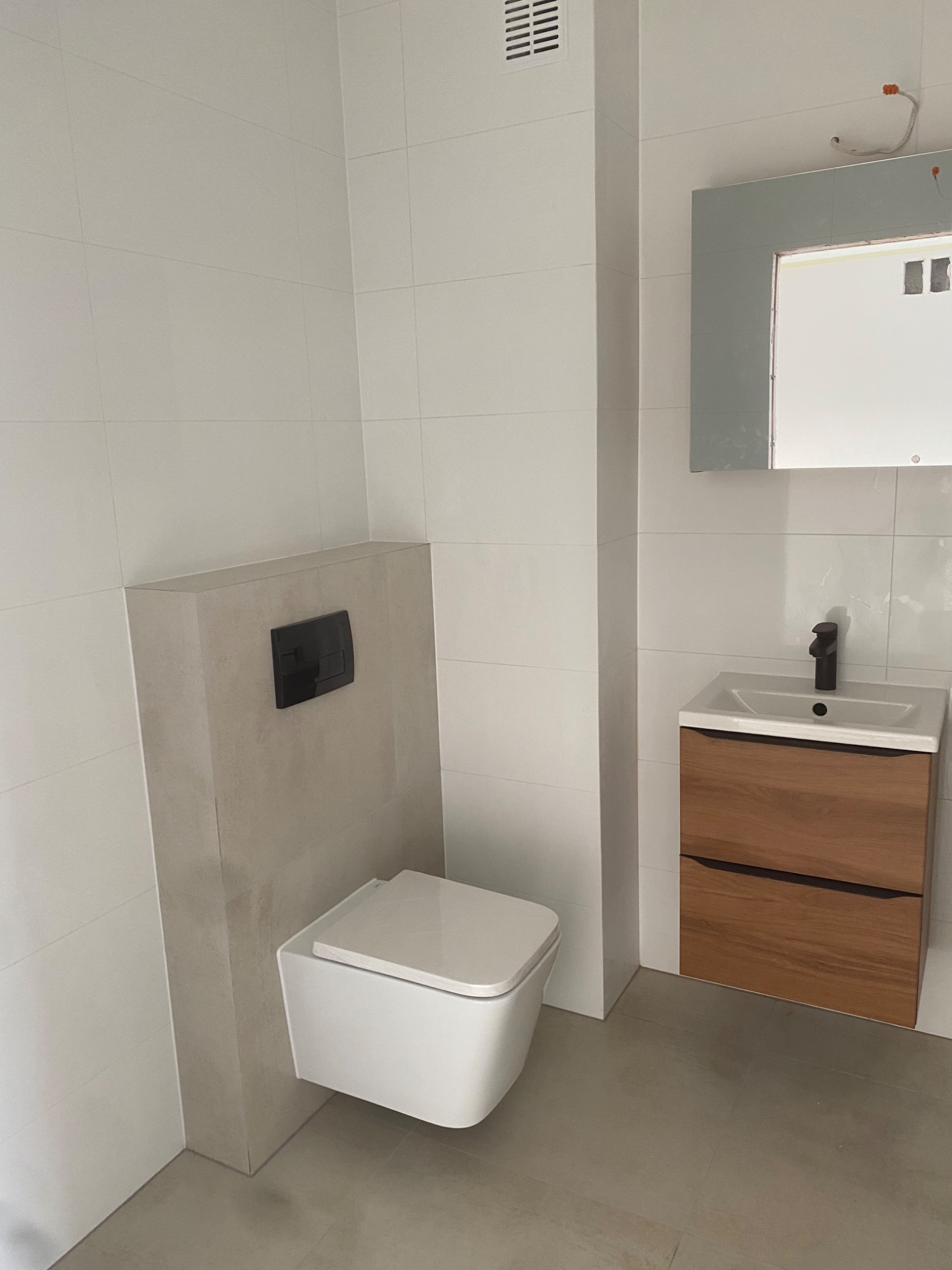 Nowoczesna łazienka z podwieszaną toaletą z czarnym przyciskiem spłukiwania, drewnianą szafką pod umywalką i dużym lustrem, wykończona białymi płytkami.
