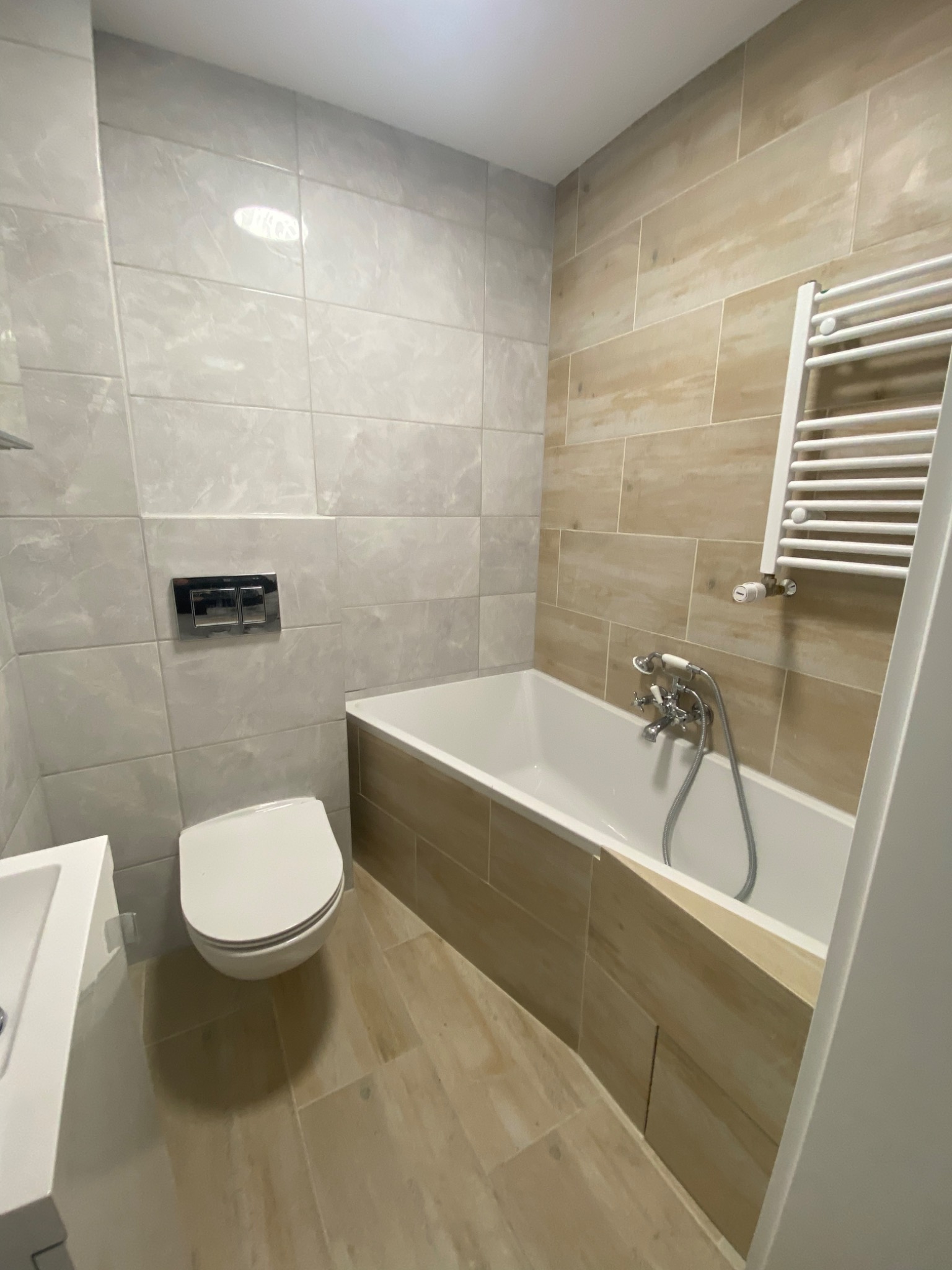 Nowoczesna łazienka z wanną obudowaną płytkami imitującymi drewno, jasnoszare płytki na ścianach, wisząca toaleta z chromowanym przyciskiem oraz biały grzejnik drabinkowy.