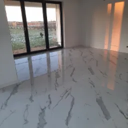 Nowoczesne wnętrze z białymi płytkami podłogowymi imitującymi marmur, duże okno z widokiem na zimowy ogród, ściany pomalowane na biało, promienie zachodzącego słońca na ścianie.