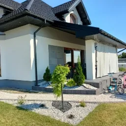 Elewacja domu jednorodzinnego po malowaniu na biało, z szarą podmurówką i czarnym dachem, widoczne elementy dekoracyjne z kamienia i zadbany ogród z rowerami.